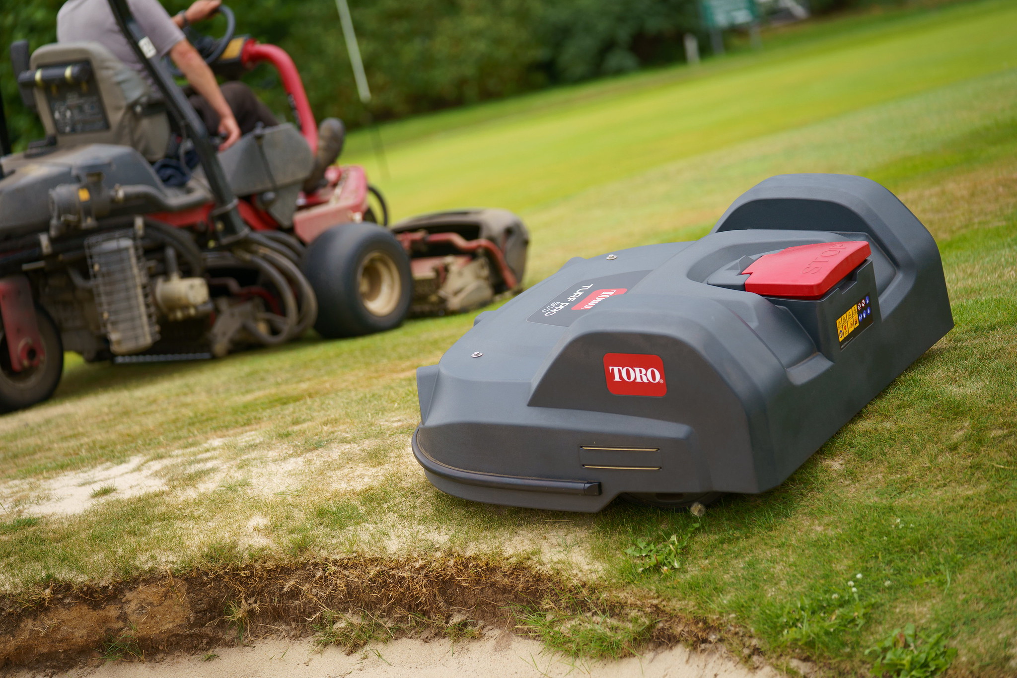 A Toro Turf Pro mows the edge of a bunker while a Toro ride-on mowers is mowing in the background