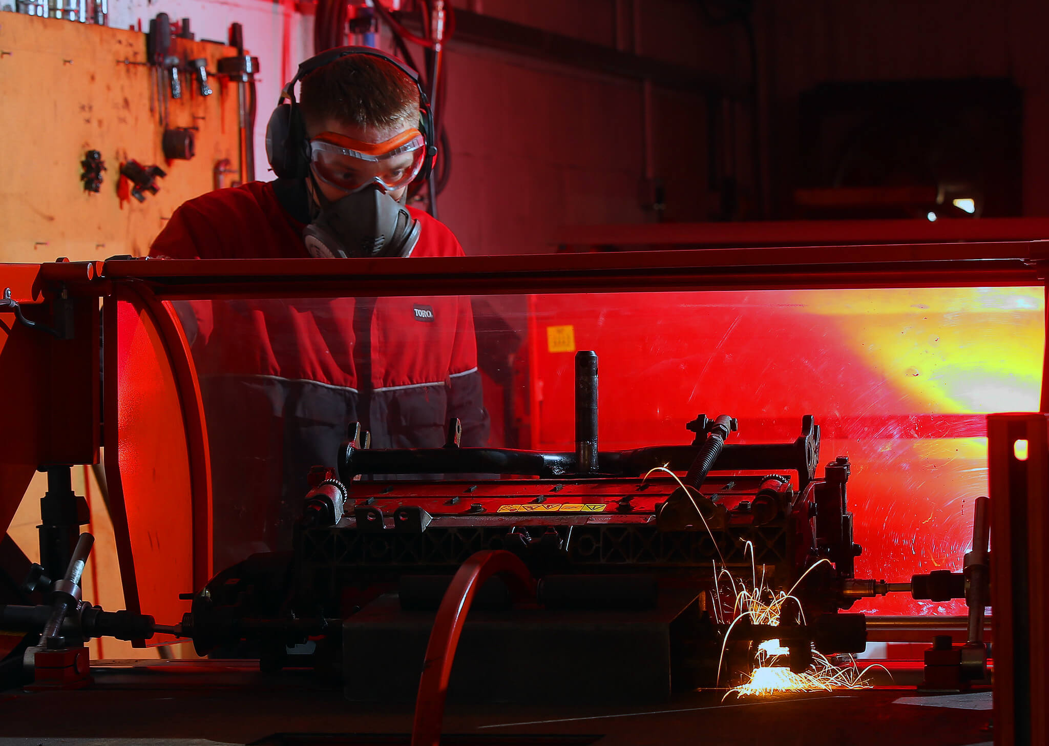Turfcare mechanic stands behind a protective screen wearing face protection as a machinery part is welded.