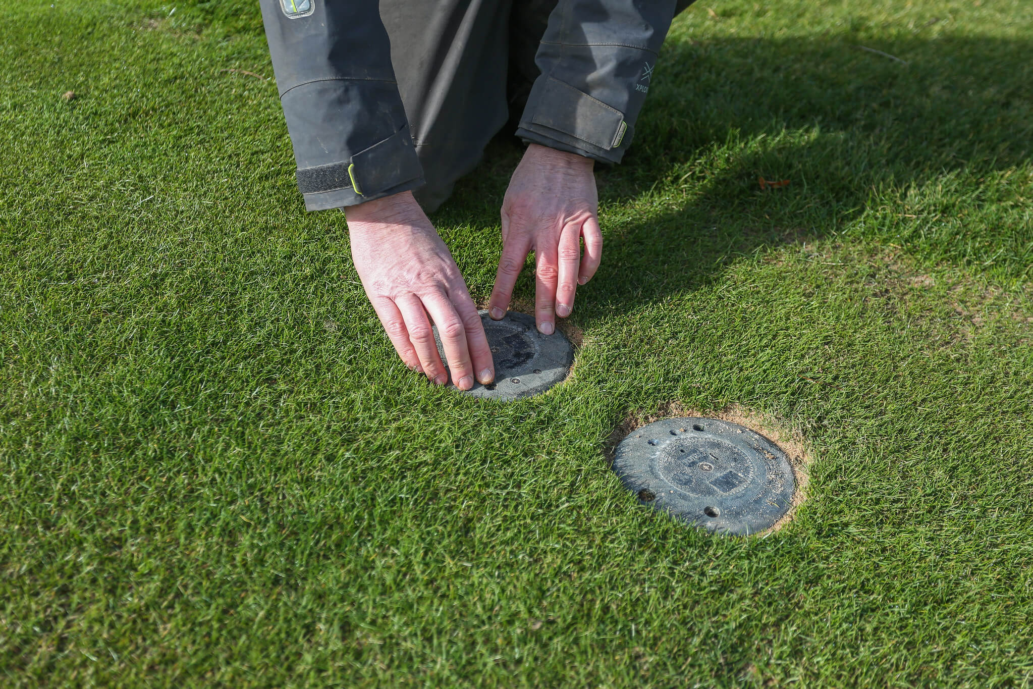 A greenkeeper adjusts two golf course sprinklers which are flush with the turf.