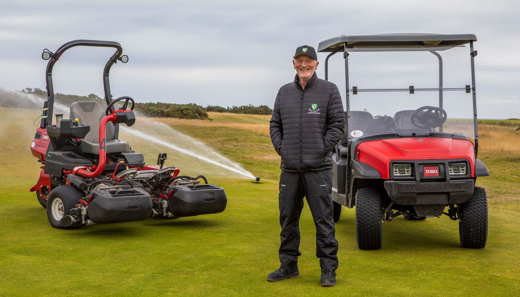 Course manager stood on the golf course with a Toro Workman, mower and sprinkler in the background