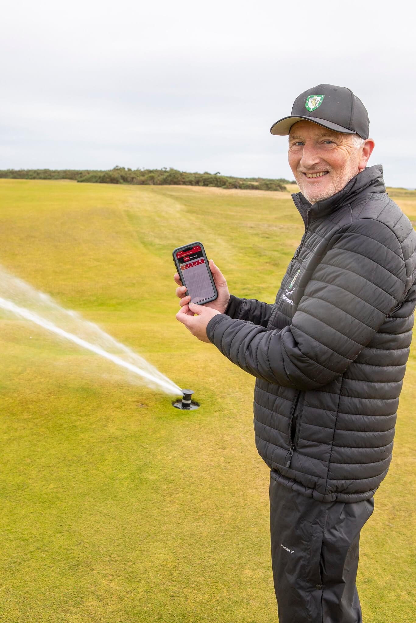 Course manager demonstrates the Lynx Central Control System on his phone while sprinkler waters in the background