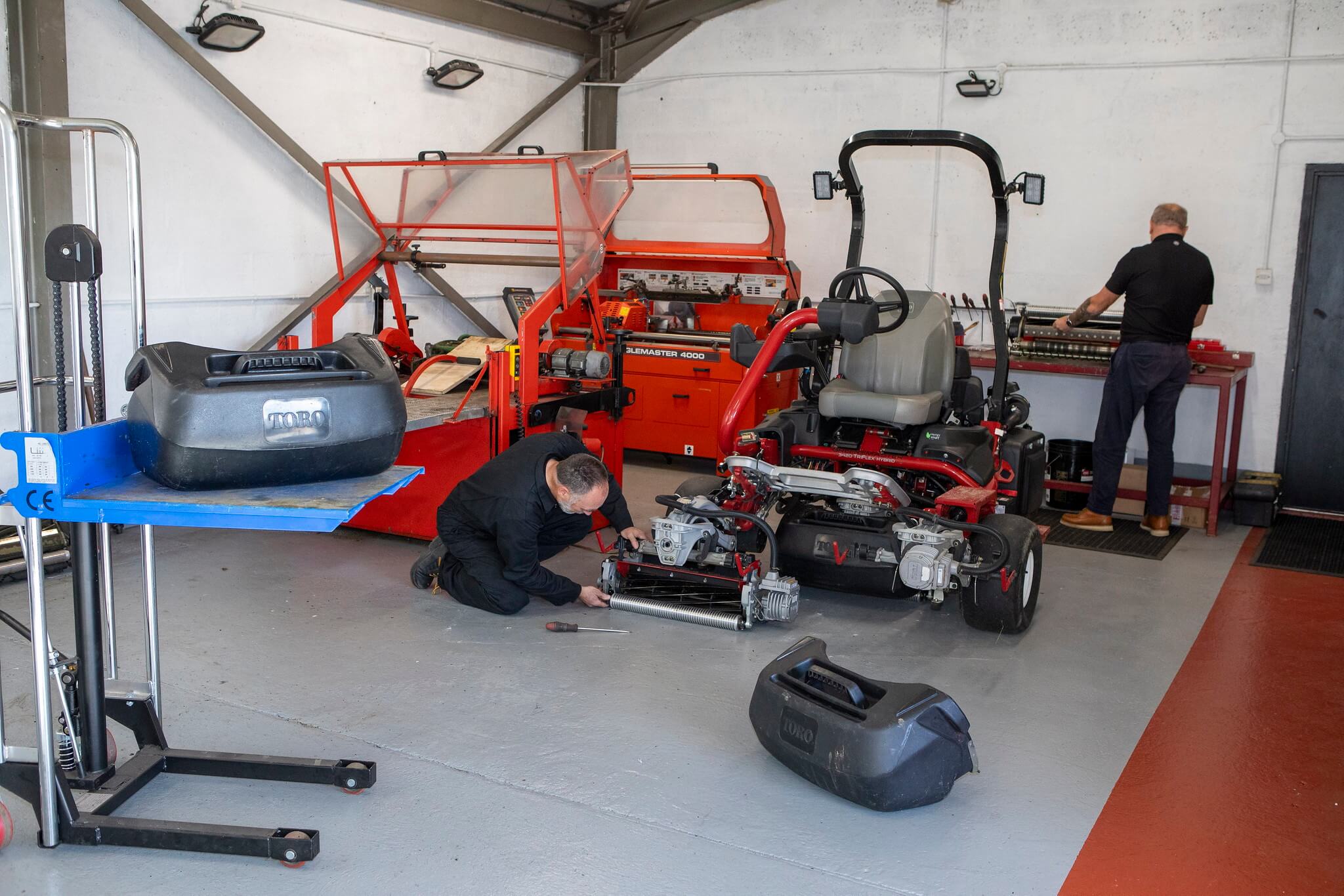 A turfcare mechanic kneels on the ground to fix a Toro mower while another mechanic stands at a workbench.