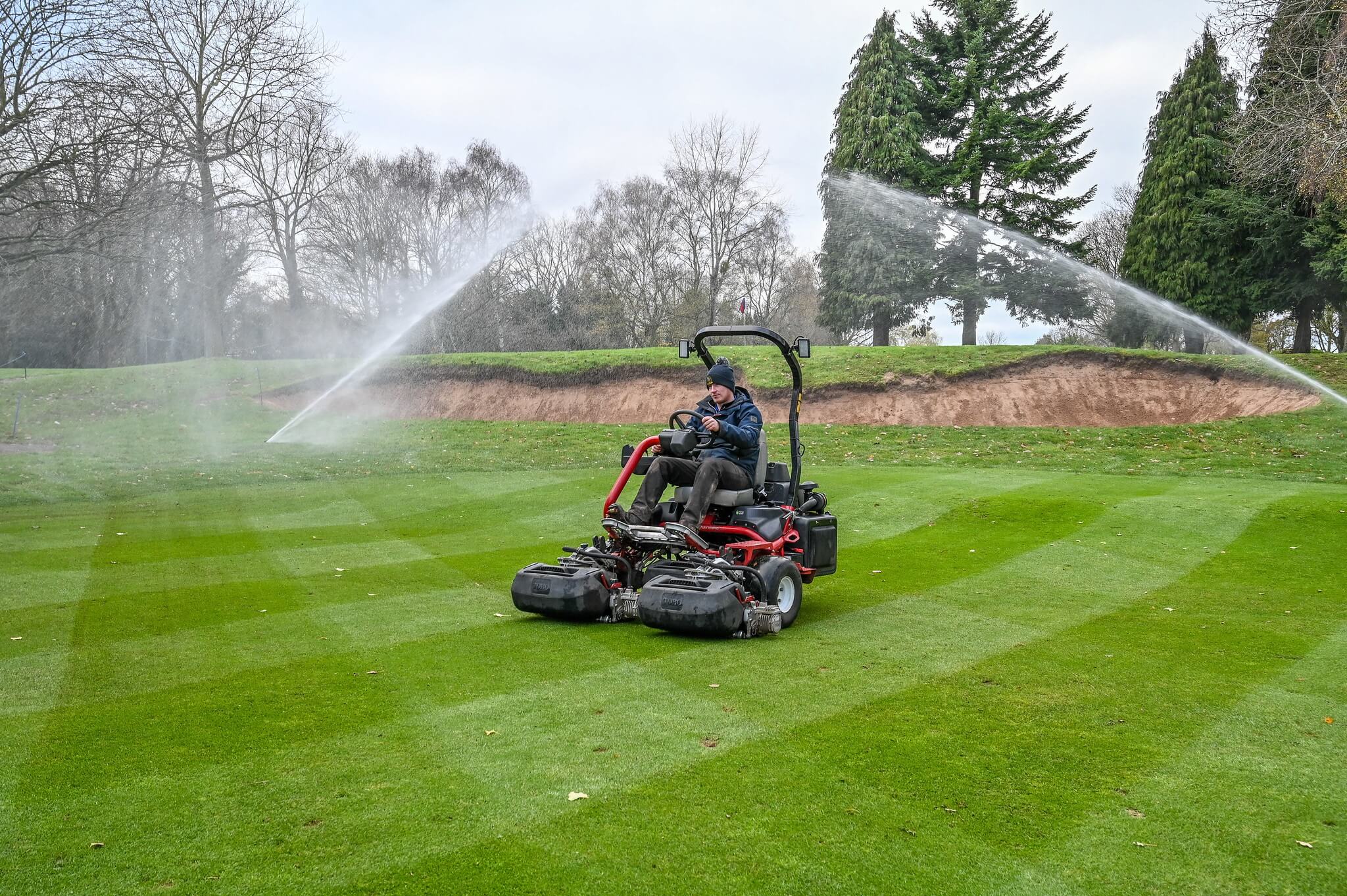Greenkeeper rides Toro mower in the fore while sprinklers are watering the course in the background