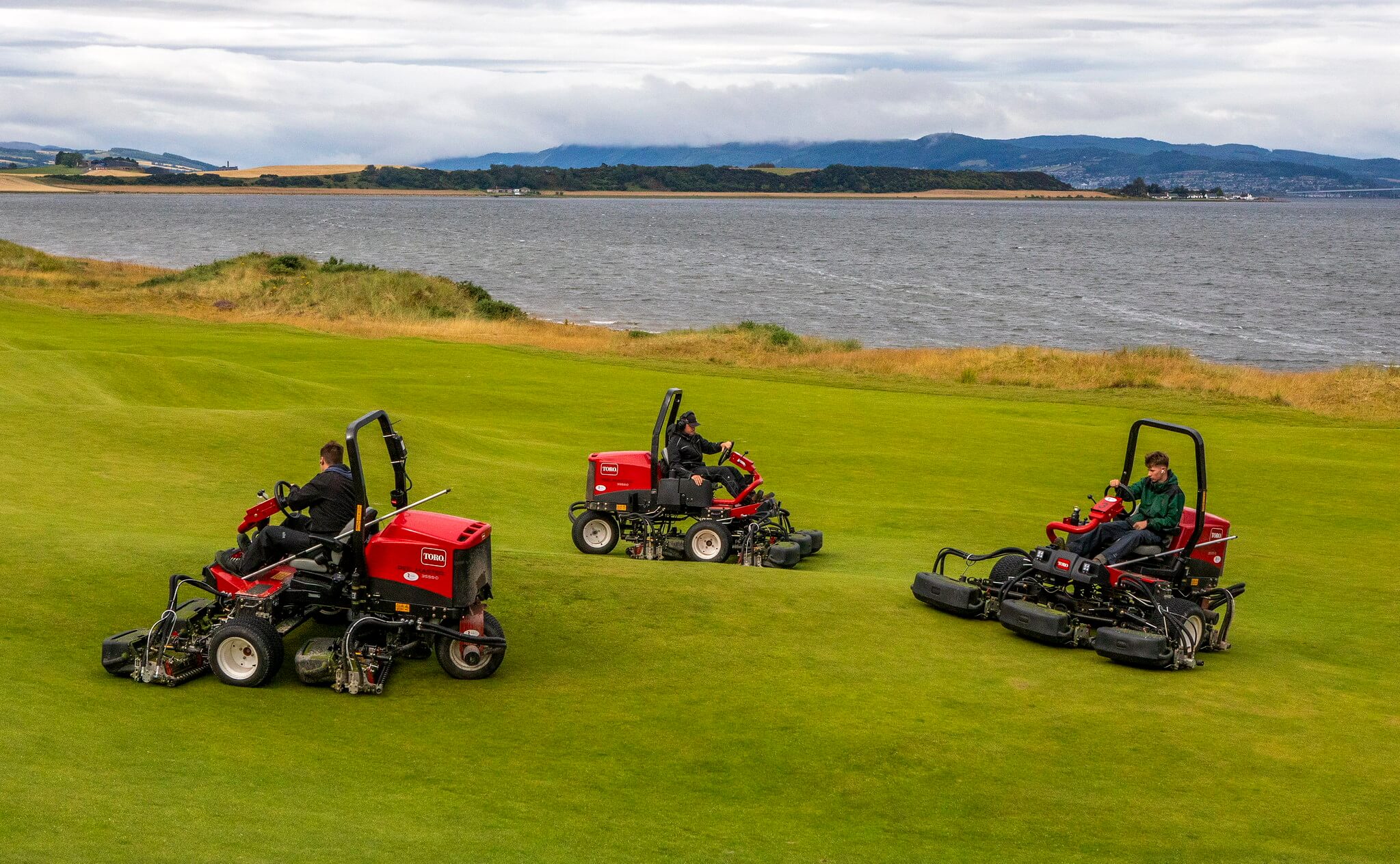 Three groundskeepers operate Toro Reelmaster mowers across a rolling coastal golf fairway at Cabot Highland