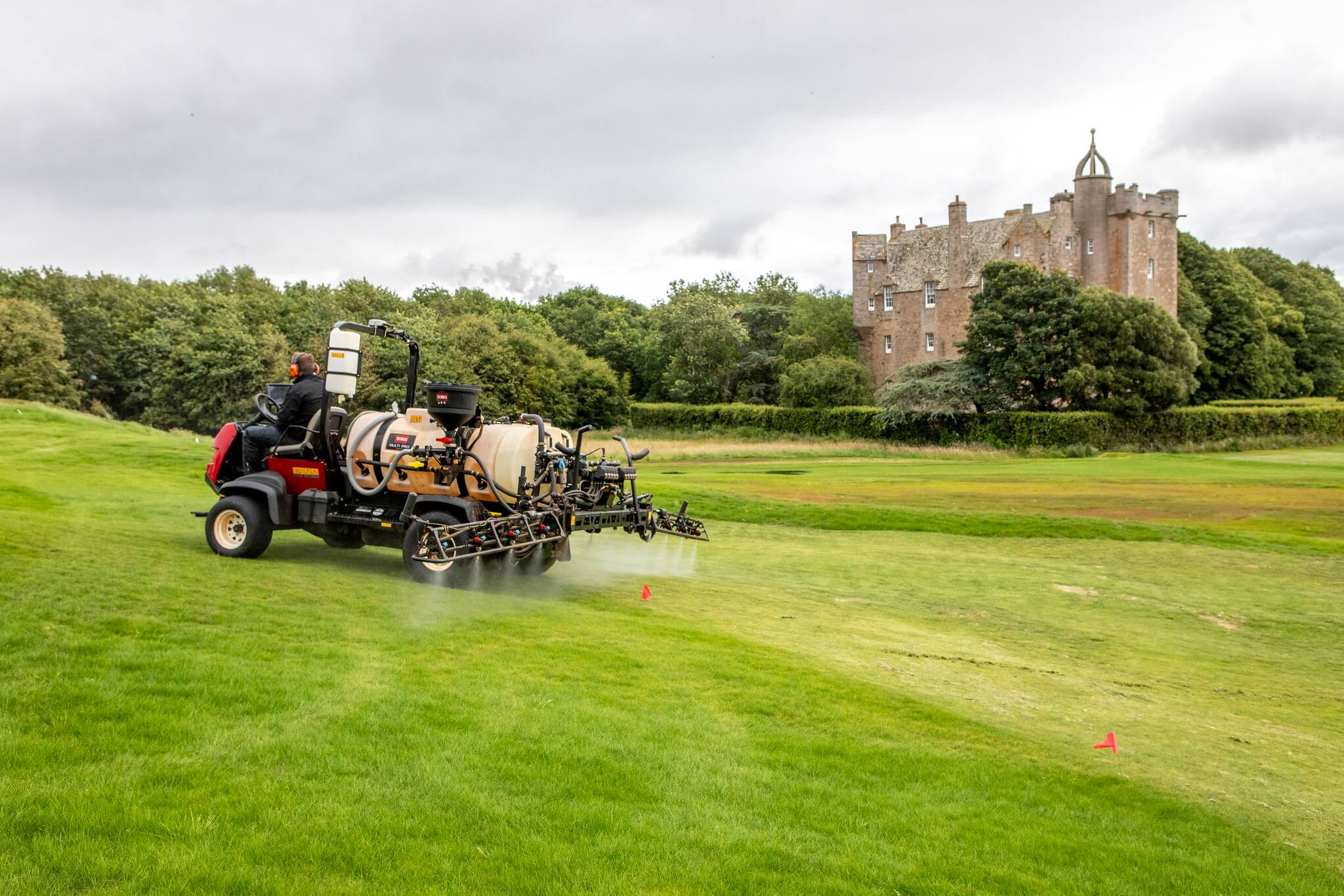 A groundskeeper drives a Toro Multi Pro sprayer across a golf course, applying treatment to the turf