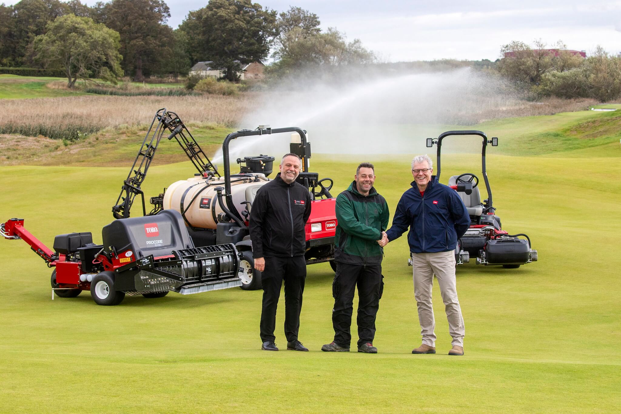 James Hutchison shaking hands with The Toro Company’s Simon Squires and Robert Jackson from Reesink Hydro-Scapes in front of Toro turf maintenance equipment, with sprinklers spraying water across the course in the background