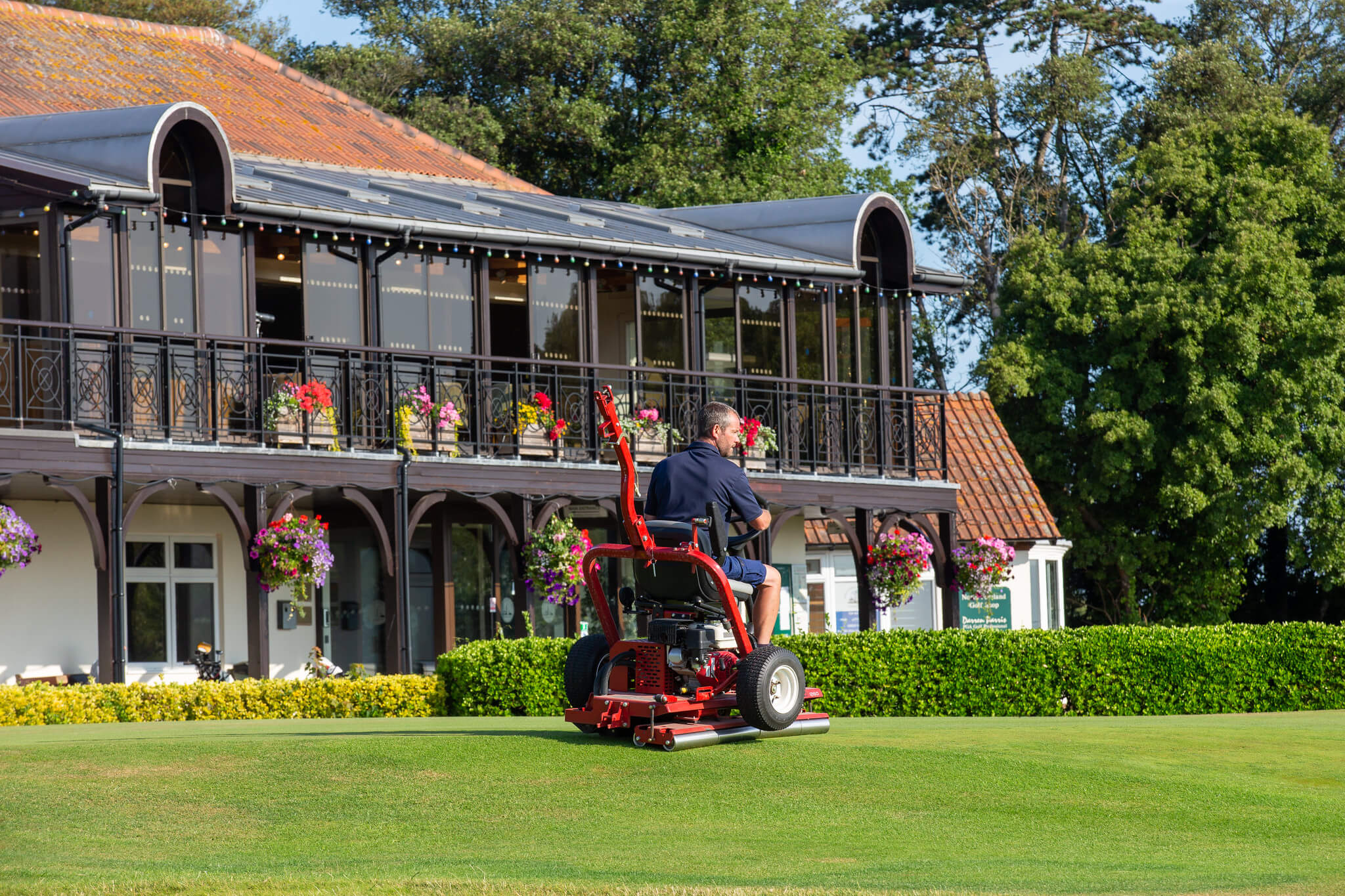 A greenkeeper rides a Toro GreensPro turf roller on a golf course with the clubhouse in the background.