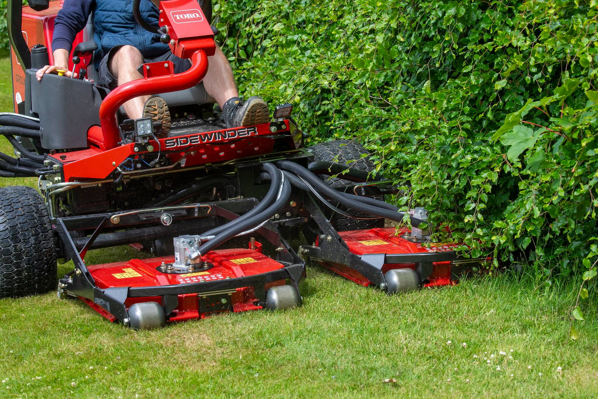 A Toro Groundsmaster mowing closely under a hedge.