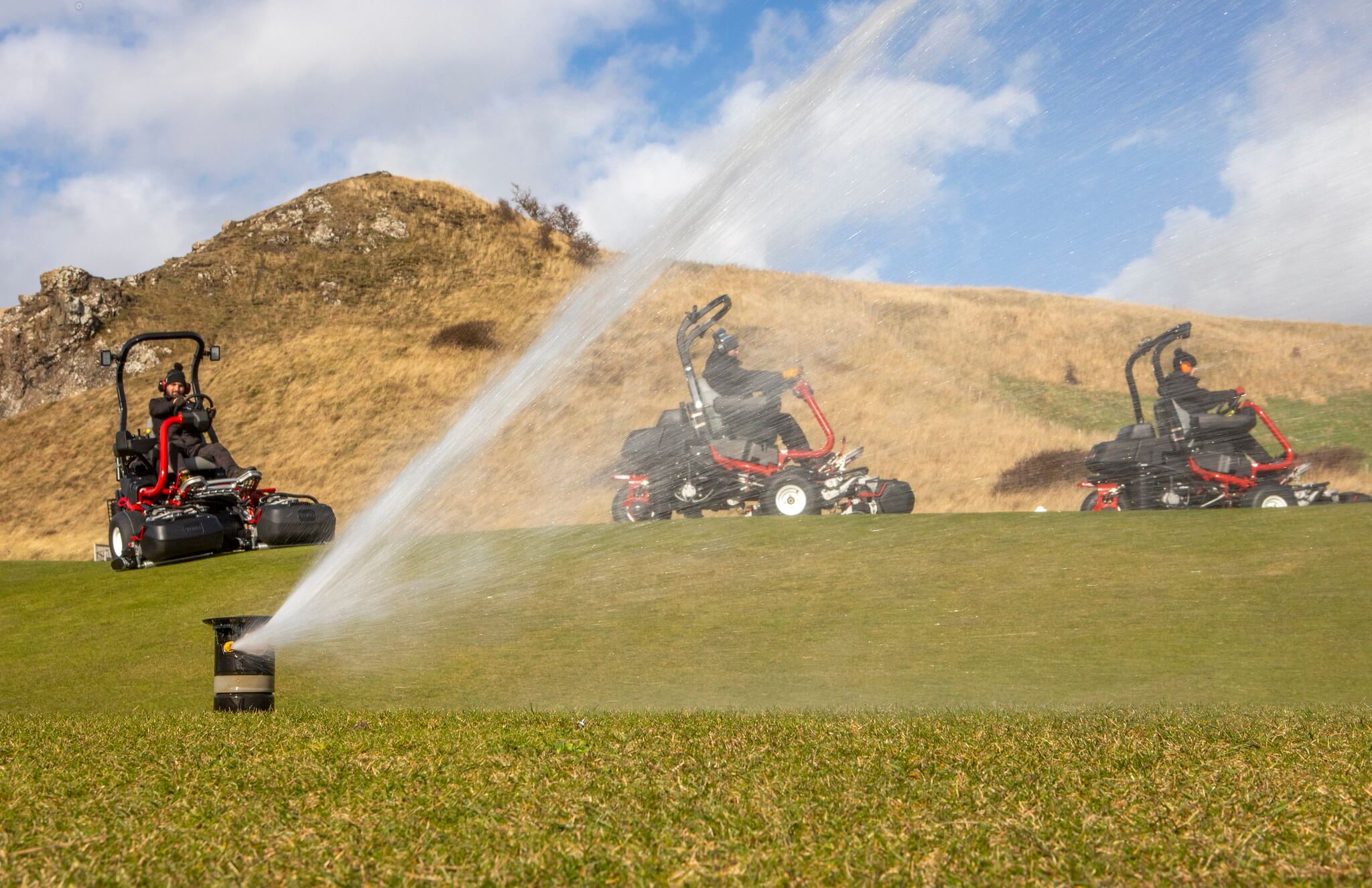 A fleet of ride-on mowers with a sprinkler in front watering a golf course.