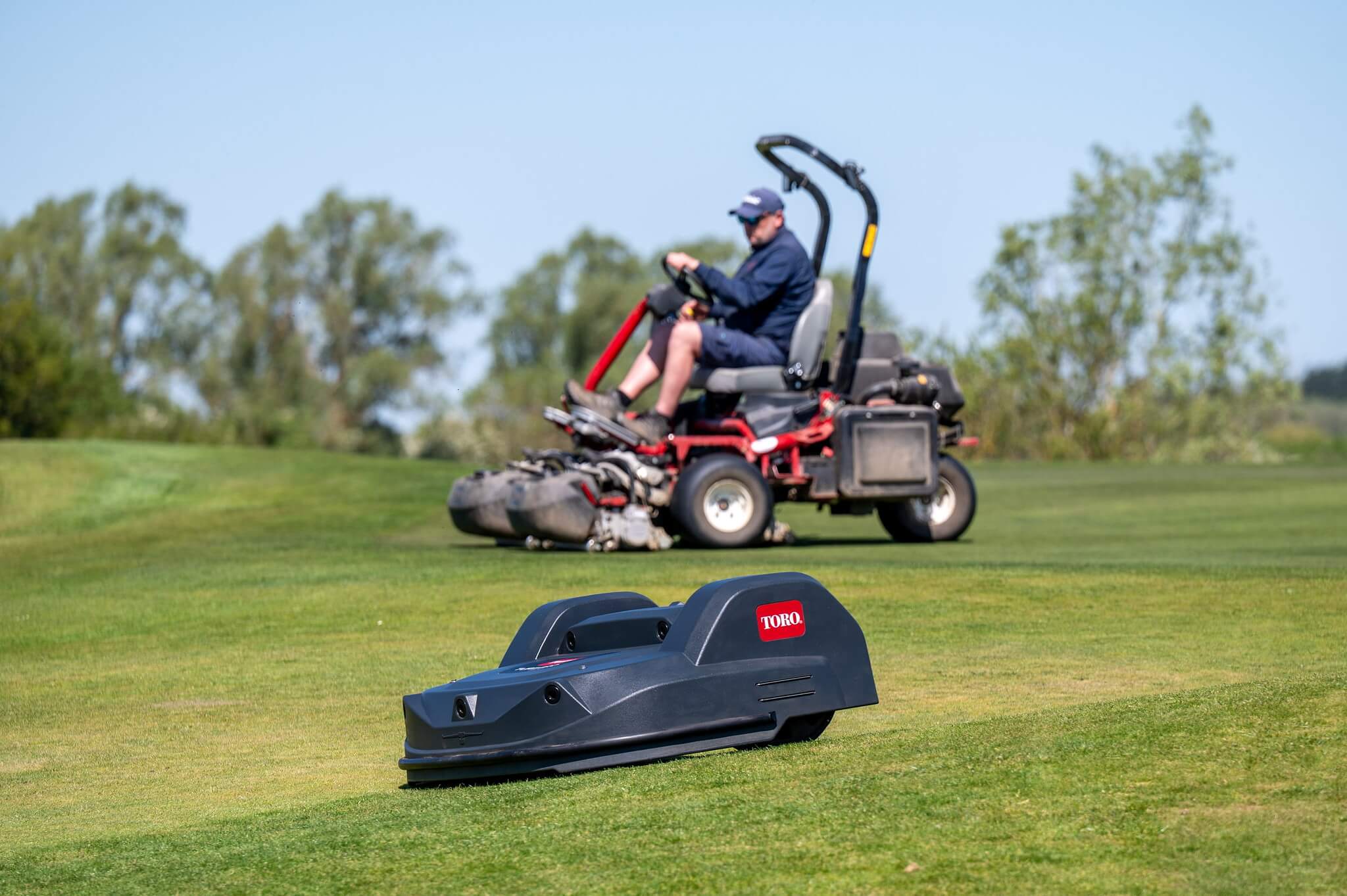 A Toro Turf Pro robot mower at work on a golf course with a greenkeeper cutting with a Toro ride-on mower in the background.
