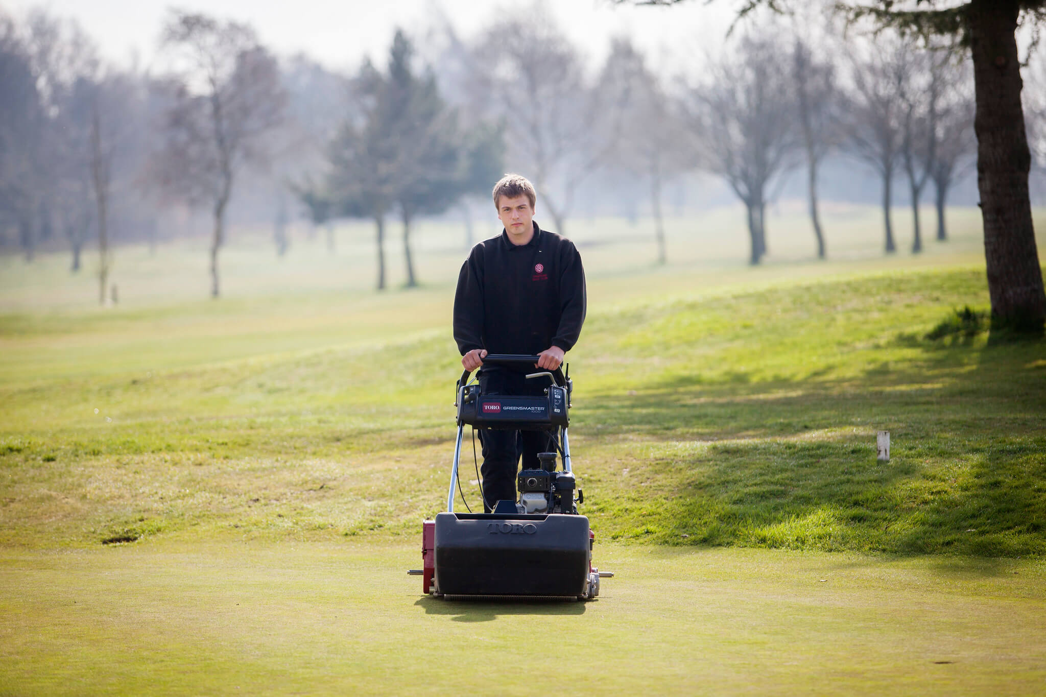 A greenkeeper using a walk behind golf mower on a course.