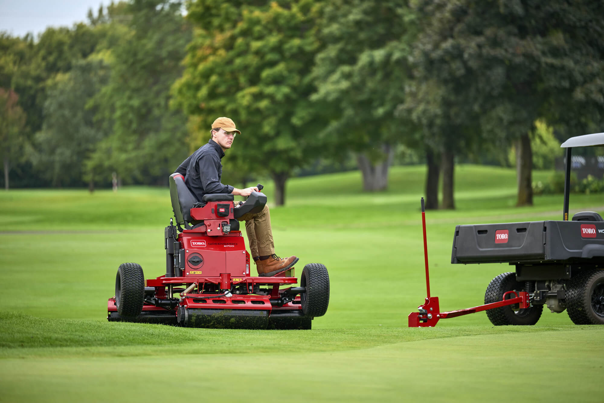 A Toro GreensPro e1700 all-electric greens roller approaching a Workman utility vehicle on a golf course.