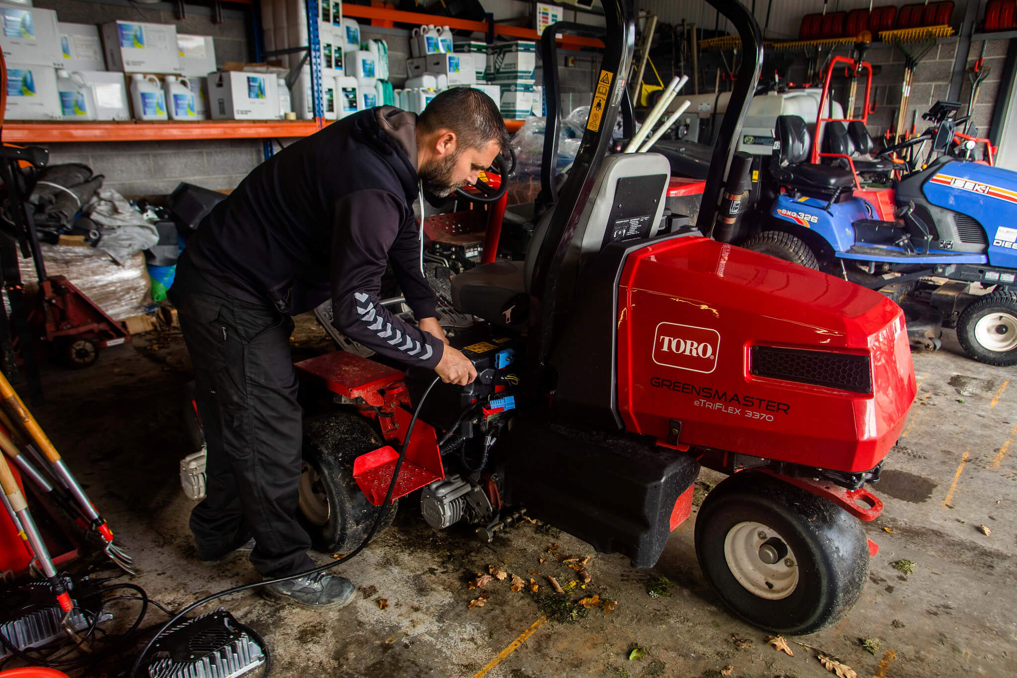 A Toro Greensmaster eTriFlex 3370 all-electric ride-on cylinder mower being charged in a machinery shed.