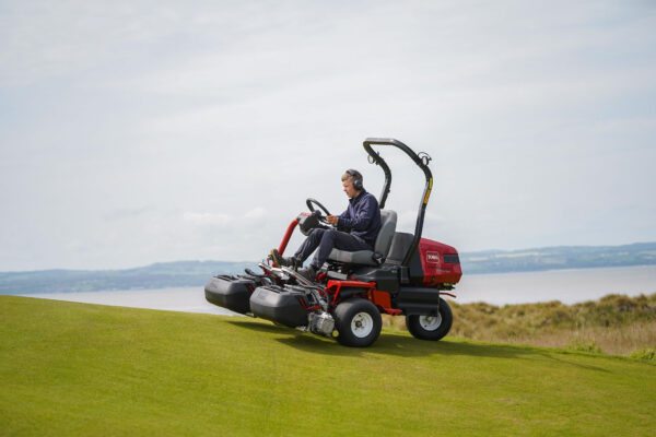 A greenkeeper driving a Toro Greensmaster eTriFlex 3370 all-electric ride-on cylinder mower on a golf course.
