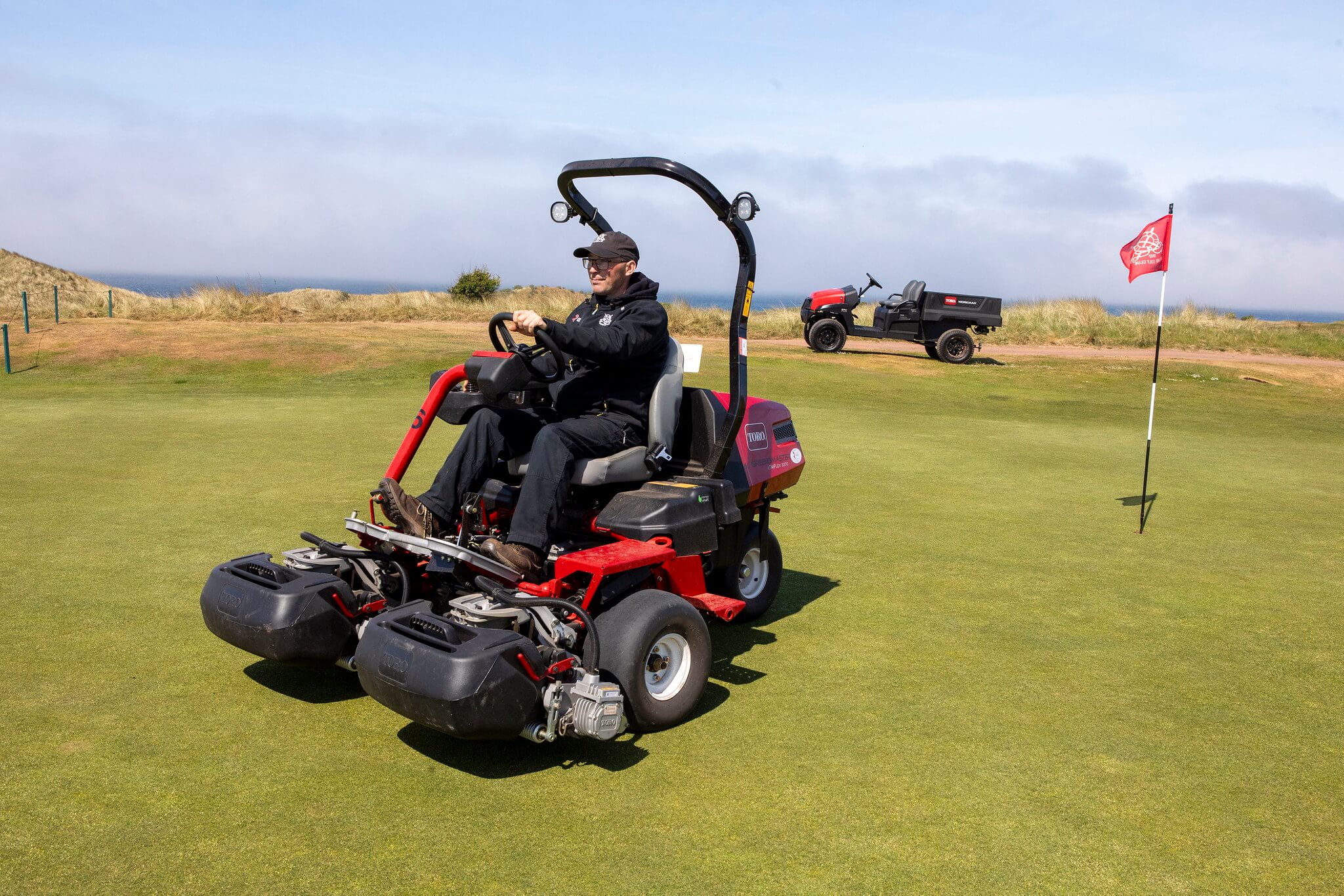A greenkeeper on a golf green driving a Toro Greensmaster eTriFlex 3370 all-electric ride-on cylinder mower with a Workman utility vehicle in the back.