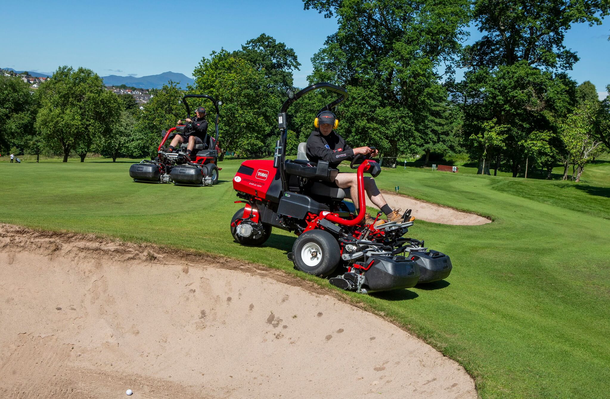 A Toro Greensmaster eTriFlex 3370 all-electric ride-on cylinder mower being ridden down a slope next to a sand bunker.