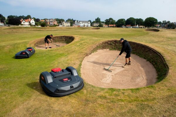 Two greenkeepers rake golf course bunkers while two Toro Turf Pro 500 robotic mowers mow around them.
