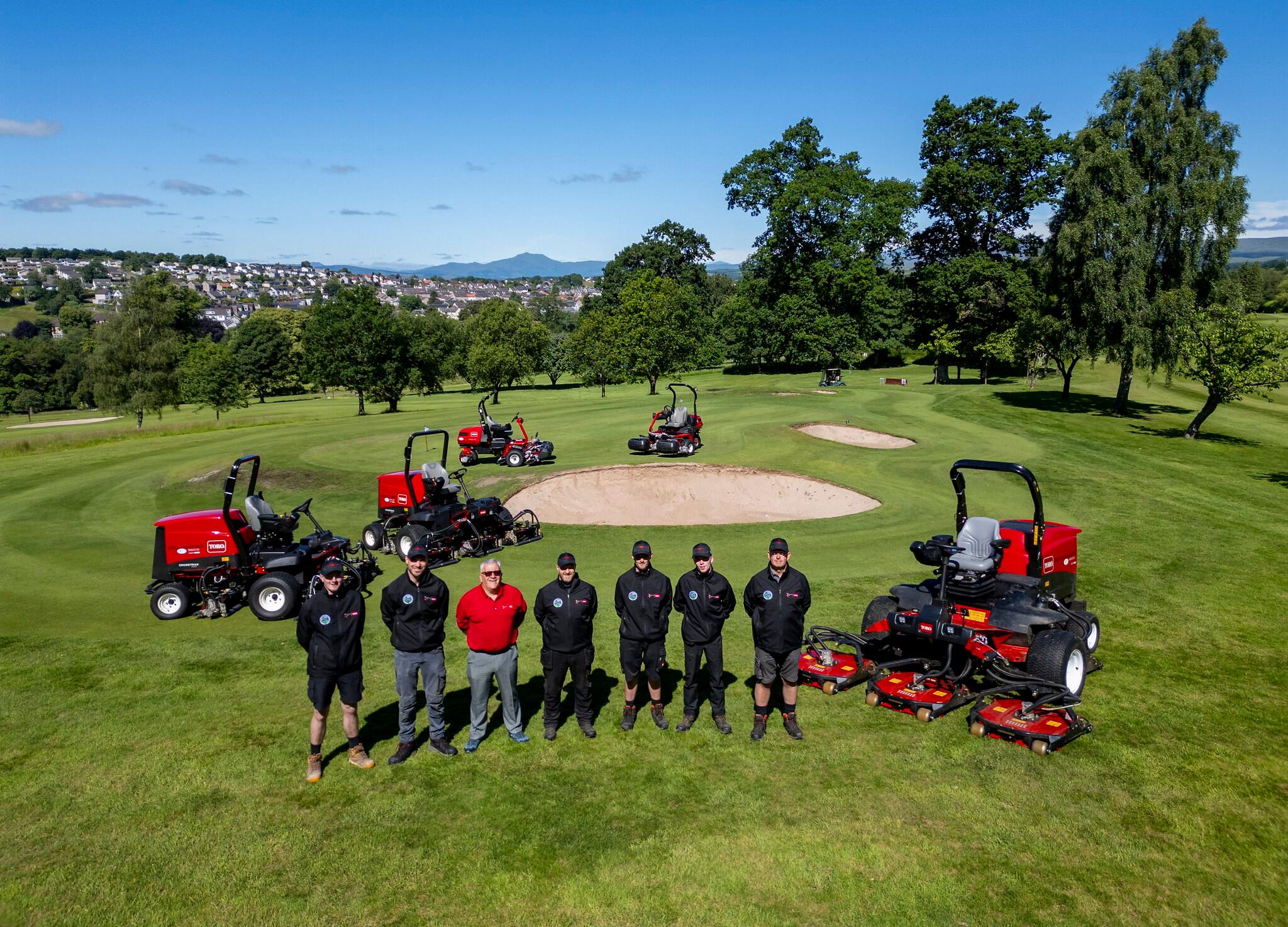 six Dunblane greenkeepers stand next to Reesink sales rep on golf course with five red Toro ride-on mowers in the background.
