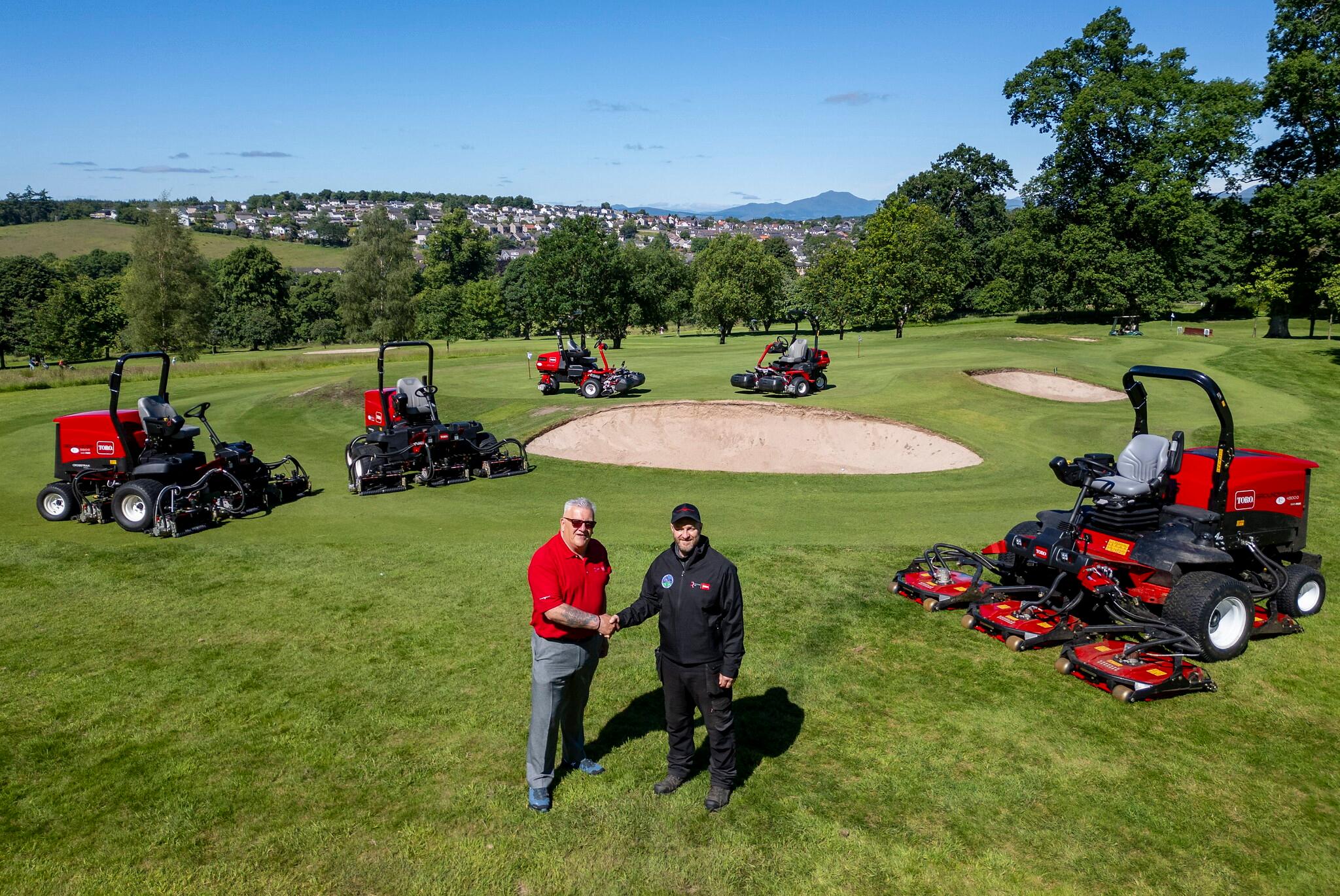 Reesink sales rep left shaking hands with Dunblane course manager right standing on golf course. There are five red Toro ride-on mowers in the background surrounding them.