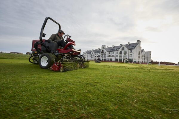 A greenkeeper sitting on a red Toro cylinder mower cutting grass on golf course with white building structure in the background.