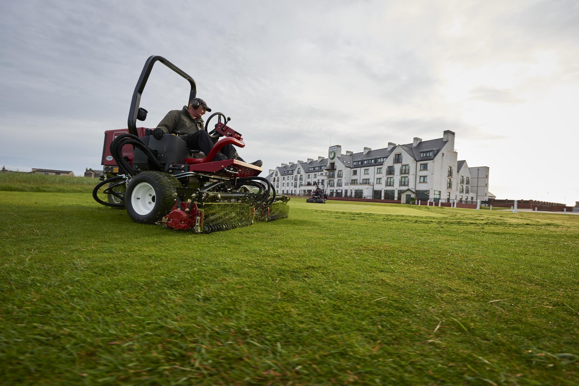 A greenkeeper sitting on a red Toro cylinder mower cutting grass on golf course with white building structure in the background.