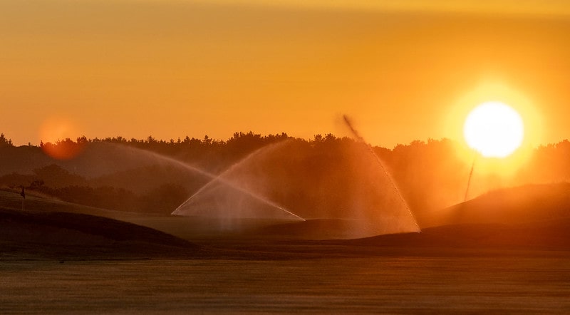 The sun rising over a series of sprinklers working on a golf course.